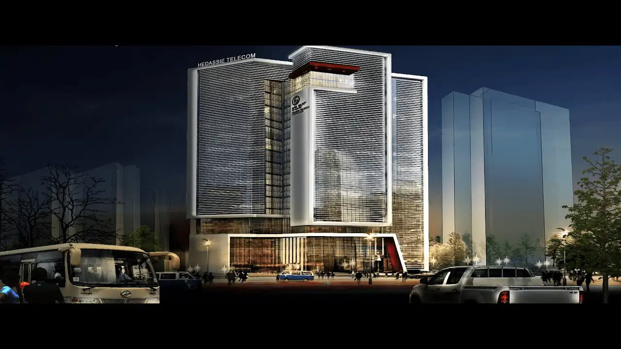 Modern multi-story office building labeled HEDASSIE TELECOM, illuminated at night with people and vehicles in front, surrounded by trees and other tall buildings, conveying a sense of progress and professionalism
